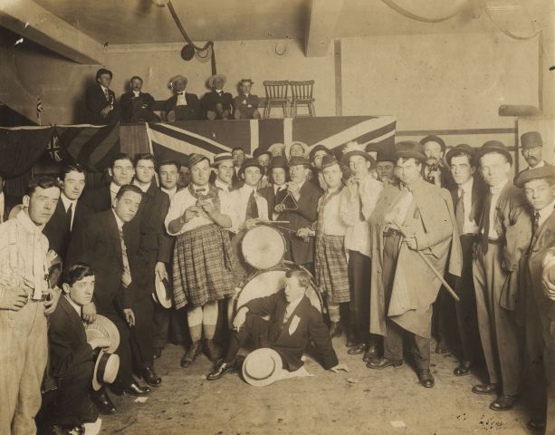 Sepia photography of a group of around thirty men, standing in a semicircle, posing for the camera. They wear suits and ties, except for two who wear kilts. In the centre, a man sits on the floor in front of a drum set. Behind the group, five men sit on a platform draped with a British flag.
