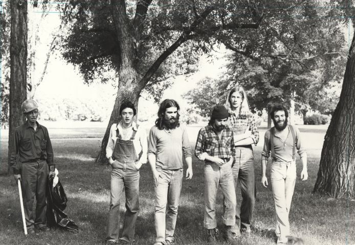 Photographie en noir et blanc d’un groupe de cinq hommes d’âge moyen, dans un parc boisé. Trois portent la barbe, deux ont les cheveux longs. Ils marchent vers la caméra, dans des vêtements décontractés. À côté d’eux, un homme d’âge mûr ramasse des ordures.