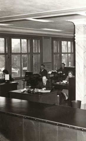 Photographie en noir et blanc de l’intérieur d’un bureau à plan ouvert. Vus de l’avant de la pièce, huit hommes et quatre femmes sont assis à des bureaux, en train de travailler. De larges fenêtres parent un des murs.