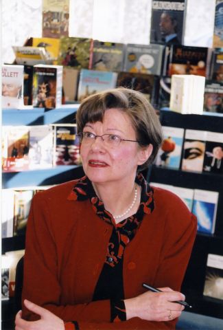 Colour photograph of a mature woman, three-quarter view. She has short brown hair, and she wears glasses, a red and black printed blouse, a red jacket, and a pearl necklace. She sits, arms crossed on a table, a pen in her right hand. In the background, a book display.