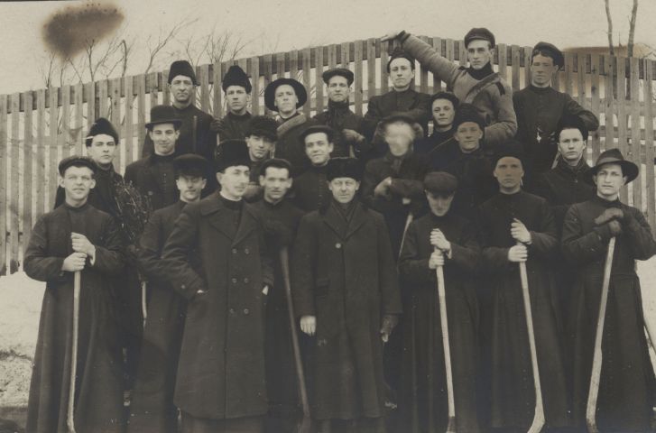 Black and white photograph, taken in winter, of a group of about fifteen young men, accompanied by some adults. They are arranged in three rows, in front of a wooden fence. The youngest wear the cassock and hold hockey sticks. Adults wear long winter coats and fur hats. One of them stands in front of the group, the other in the center of the photo, the other two on the sides.