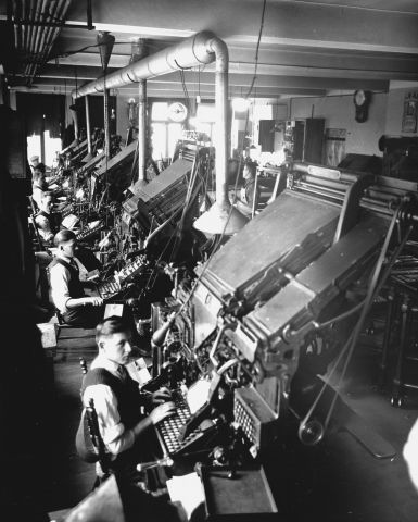 Photographie en noir et blanc de cinq hommes en chemise, gilet et cravate dans un atelier industriel. Ils sont assis devant d’imposantes machines de composition, avec clavier.