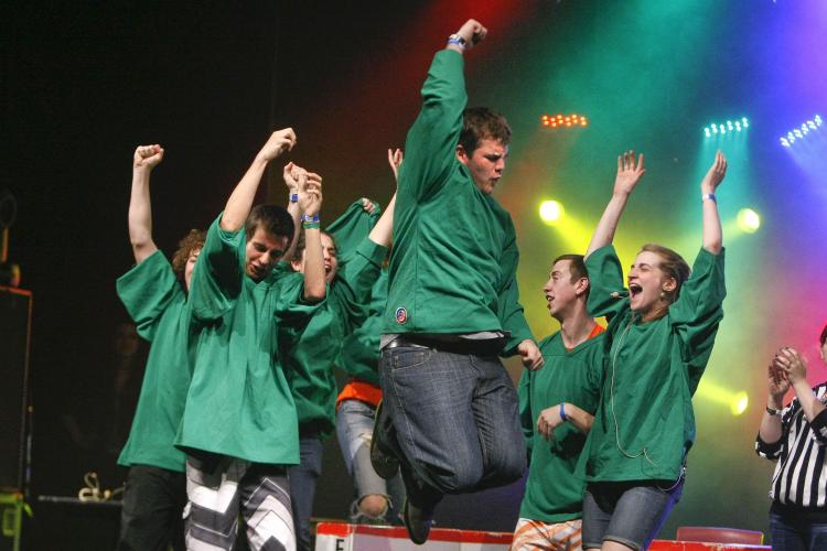 Colour photograph of a group of seven teens wearing green jerseys, celebrating a victory on stage. The young man in the foreground of the photo is jumping with joy, his right fist raised over his head.