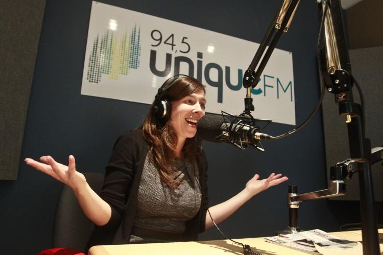 Colour photograph of a middle-aged, smiling woman in a radio studio. She wears headphones and speaks into a suspended microphone. She sits at a table with her arms raised, hands open, palms up. Behind her, a sign with the words “94,5 Unique FM.”