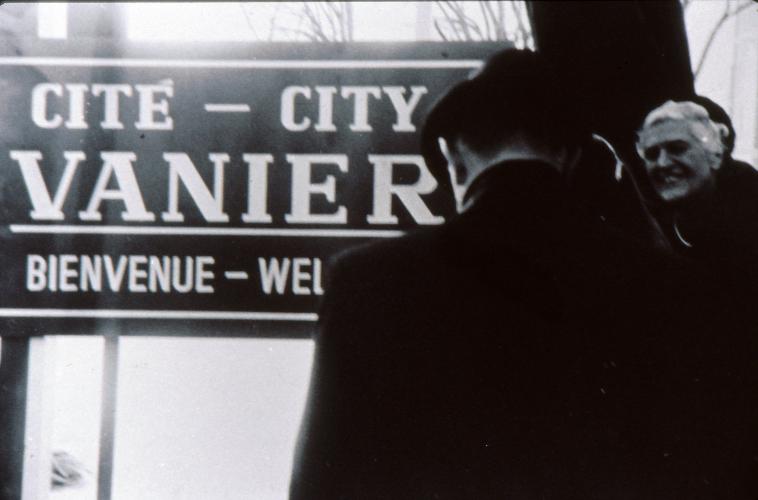 Black and white photograph showing a smiling mature woman and a man, from behind. Both are dressed with black coats and wear hats. They stand in front of a bilingual Vanier welcome sign.
