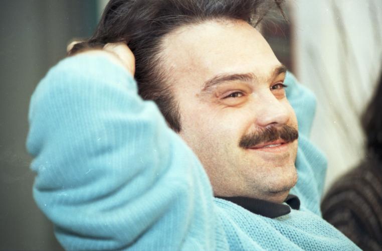 Colour photograph of a middle-aged man, three-quarter-view. He is smiling, sitting with both hands behind his head. He has brown hair and a mustache. He wears a pale blue sweater.