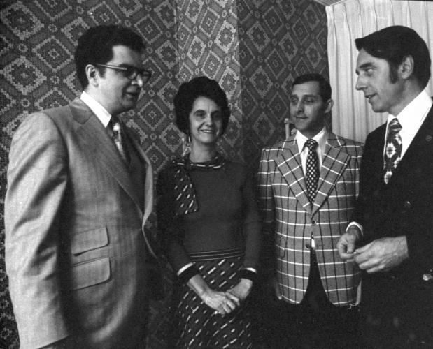 Black and white photograph of three men and a woman, middle-aged, talking. They are standing in a living room. The men wear jackets and ties; the woman wears a dress.