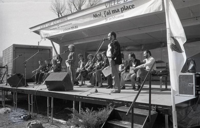 Black and white photograph of a middle-aged man and woman standing at microphones on an outdoor stage. Behind them, around ten people sitting on benches. Written on a banner above the stage: “Centre national des Arts–National Arts Centre; Théâtre français saison 83-84; Moi, j’ai ma place; Je m’abonne.”