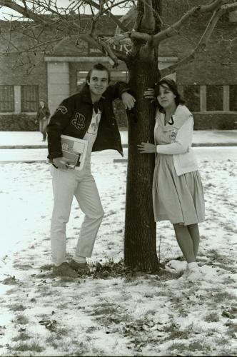 Black and white photograph of two teenagers, male and female, outside a brick building in winter. They lean casually against a tree. He wears a leather jacket and a shirt with a fleur-de-lis; she wears a dress with a large collar and a white jacket.