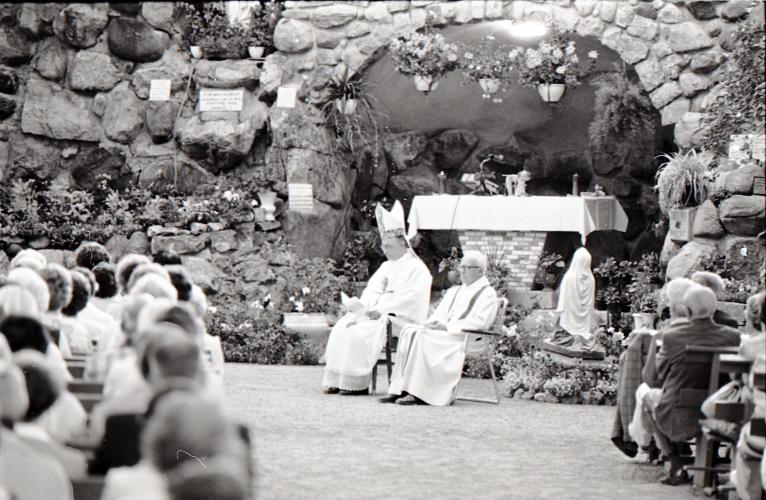 Photographie en noir et blanc d’un évêque et d’un prêtre vêtus de blanc assis devant une grotte décorée de fleurs et d'une effigie de la Vierge. De nombreux fidèles, vus de dos, sont assis sur de longs bancs ordonnés en rangées.