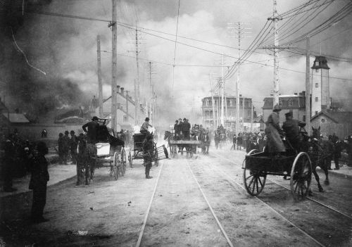 Black and white photograph of a large crowd in a street threatened by smoke. Some people are in buggies with their possessions. Firefighters arrive in wagons.