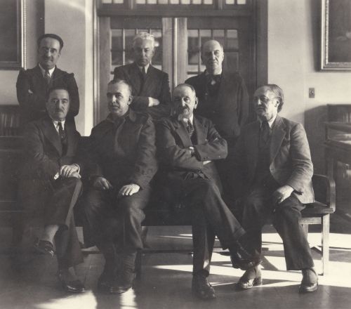 Black and white photograph showing seven older men in an exhibition hall. Four sit on a bench, while three stand behind them.  They are all dressed in suits and look serious.