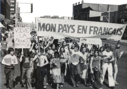 Black and white photograph of a group of young people protesting in the street. Individual at the front carry a banner reading “Mon pays en français“ and a placard reading “Vive la francophonie“.