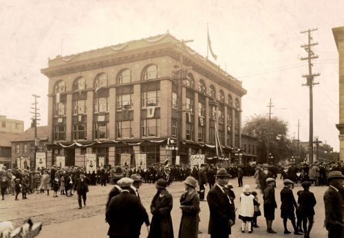 Black and white photograph of a large crowd, on both sides of a city street. Men, women and children wear elegant clothes. In the background, a four-storey building, richly decorated.Many banners of the Virgin Mary and other patron saints.
