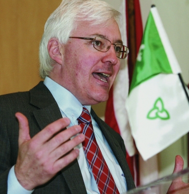 Colour photograph of a mature man with white hair and glasses, in a dark gray suit, white shirt, and red-patterned tie. He is standing in front of a Franco-Ontarian flag, delivering a speech.