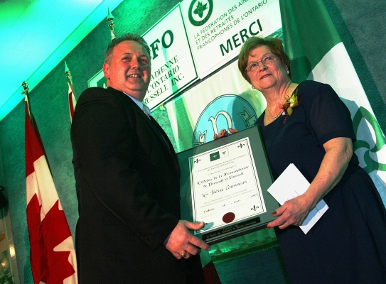 Colour photograph, slightly green-tinted, of a middle-aged man and an older woman standing on a stage, seen from below. They are holding a framed certificate between them, and smiling lightly. In the background, mostly green, a Canadian flag, Franco-Ontarian flag, and the logos of various organizations.