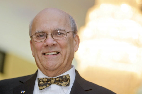 Close-up colour photograph of an older, balding man wearing glasses, smiling at the camera. He wears a dark gray suit, white shirt and gray-gold bow tie.