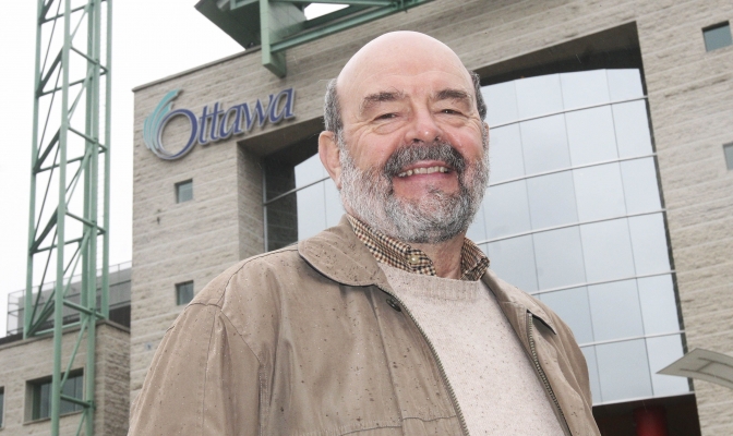 Colour photograph of an older, balding man with a salt and pepper beard, smiling at the camera. He wears a beige coat splattered with raindrops, and stands in front of a multi-storey, concrete and glass building bearing the City of Ottawa sign.