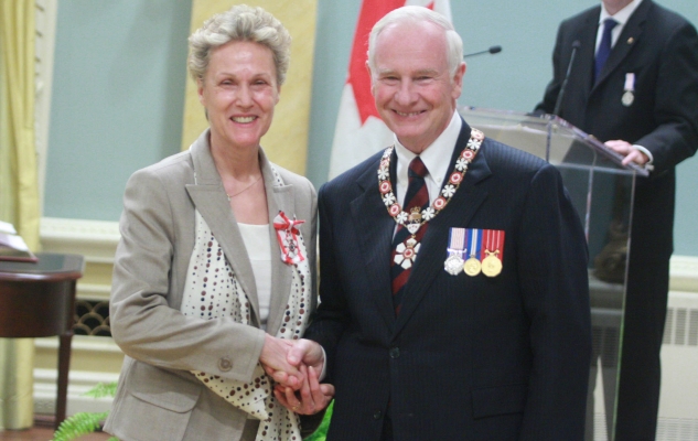 Colour photograph of a mature woman wearing a beige jacket and matching patterned scarf. The Order of Canada badge is pinned to her jacket. The woman is accompanied by an elderly man wearing a suit, tie, medals and the chain of office of the Governor General of Canada. The two decorated people shake hands during a ceremony. The flag of Canada is visible behind them.