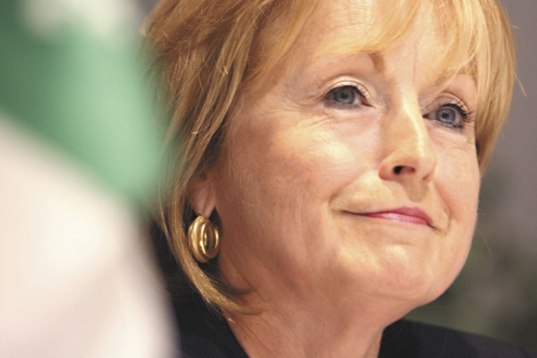 Close-up colour photograph of a mature woman with strawberry blond hair, in three-quarter view. She wears gold earrings, a dark top, and a small smile. In the foreground, part of the Franco-Ontarian flag, out of focus.