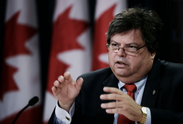Colour photograph of a middle-aged man with dark hair and plump face, wearing glasses. He is talking and gesticulating in front of a microphone. Three Canadian flags are visible behind him.