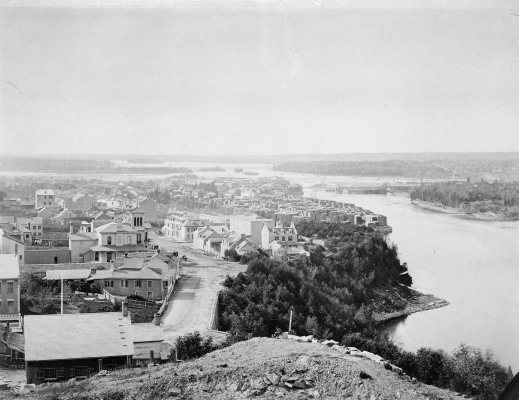 Black and white photograph of an urban landscape, viewed from above. An imposing river borders it to the right, crossed by a bridge that leads to a wooded area. In the distance, reaching far into the distance, the forest.