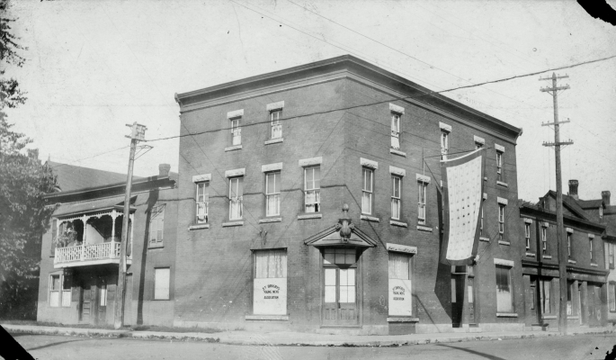 Black and white photograph of a three-storey brick building. Posters in two windows on the ground floor bear the name of the organization. A banner patterned with maple leaves is suspended, flag-like, from the second floor of the building.