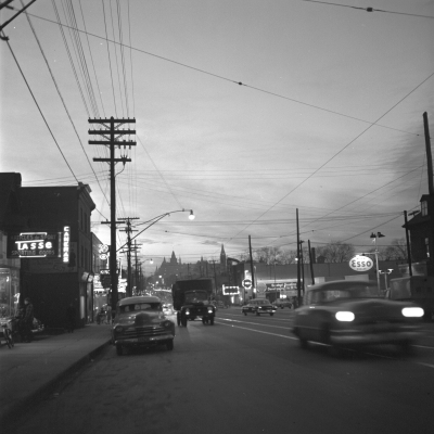 Black and white photograph of a boulevard at night. Car headlights and shop signs illuminate the photograph.