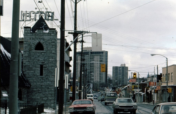 Colour photograph of a busy street with many cars. The street is lined with shops. Large buildings appear in the distance.