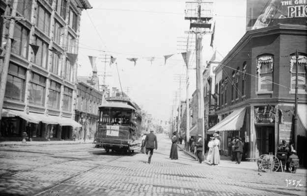 Black and white photograph of a streetcar on a wide commercial boulevard decorated with streamers. People walk on the sidewalks and enter the streetcar.