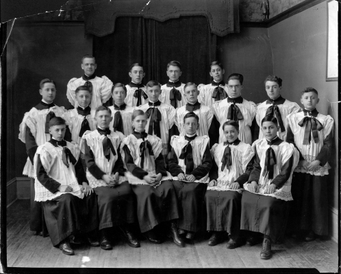 Photographie en noir et blanc, d’un groupe d’une quinzaine de jeunes garçons. Certains sont assis, les autres sont debout derrière, en deux rangées. Tous portent des soutanes et des surplis.