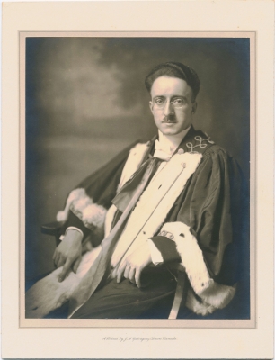 Black and white studio photograph of a serious-looking young man with glasses, dark hair, and a small mustache. He is seated and wears a fur-trimmed academic robe. The photographer’s name and studio address are printed in English at the bottom of the photograph.