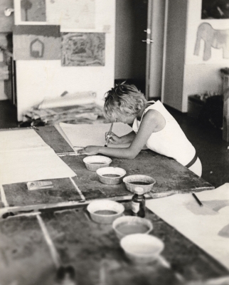 Photographie en noir et blanc d’une jeune femme dans un studio d’art. Elle est penchée sur une table recouverte de grandes feuilles de papier et de petits récipients de peinture, en train de peindre.