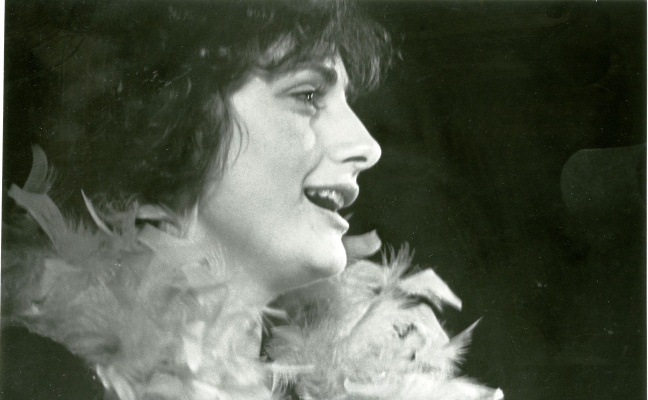 Black and white close-up photograph of a young woman in profile, singing into a microphone. She has curly, dark hair, and a feather boa around her neck.