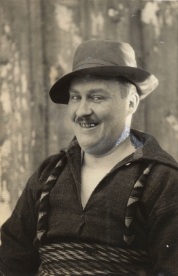 Black and white photograph of a mature man wearing a mustache, heavy shirt, hat, and woven sash. He is smiling.