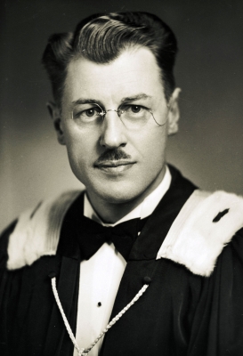 Black and white, head-and-shoulders studio photograph of a dark-haired, middle-aged man, seen from the front. He wears glasses and a small mustache. The man is dressed in a white shirt, bow tie and fur-trimmed academic robe.