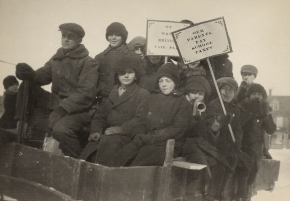 Black and white photograph of a dozen boys dressed for winter, crowded into a cart. Two of the boys brandish placards with “OUR PARENTS PAY SCHOOLS TAXES” and “WE WANT BRITISH FAIR PLAY.” Others play small musical instruments.