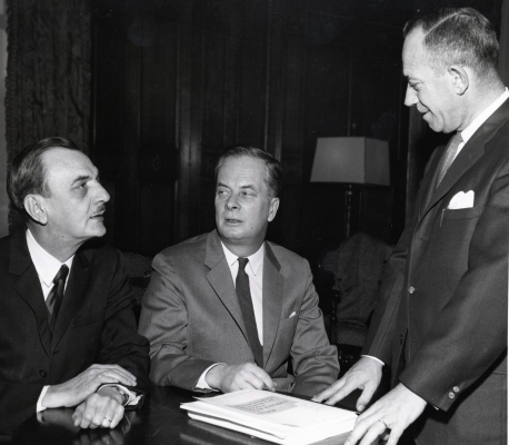 Photographie en noir et blanc de trois hommes d’âge mûr en costume-cravate. Deux des hommes sont assis côte à côte à un bureau ; le troisième leur fait face, debout. Seul un des hommes est vu de face. Les deux autres sont présentés de profil.