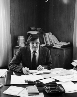 Black and white photograph of a dark-haired young man working at a messy desk, smiling at the camera. He wears a white shirt, dark suit, and dark tie.