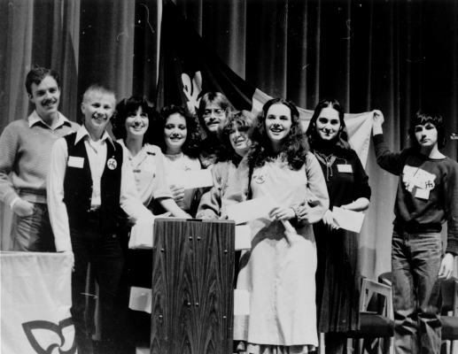 Black and white photograph of a group of young people, accompanied by some adults, grouped around a lectern. A young woman holds a document, while others hold envelopes. In the back, another group member holds up a Franco-Ontarian flag.