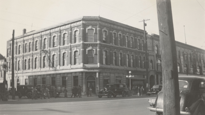 Black and white photograph of a three-story brick building with many windows. It is located on a busy street. Engraved in stone, above the main entrance and on the sides, the Bank of New Scotia name.