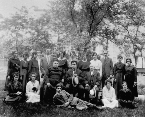 Black and white photograph of a group of twenty people, mostly young women. Three older men, including a priest, are seated at the center of the image. A young man is reclining on his side in front of them. The others are standing or sitting on the ground. The photograph was taken outside.