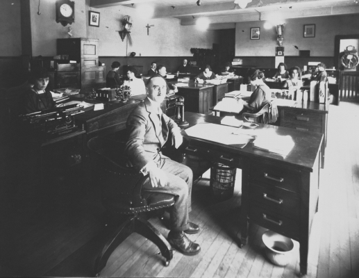 Black and white photograph of men and women working at their desks. In the foreground, a middle-aged man in a suit looks at the camera. To his left, a serious-looking young woman behind her desk.