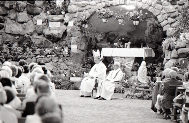 Black and white photograph of a bishop and a priest dressed in white, sitting before a cave decorated with flowers and an effigy of the Virgin. Many of the faithful, seen from behind, are seated on long benches arranged in rows.