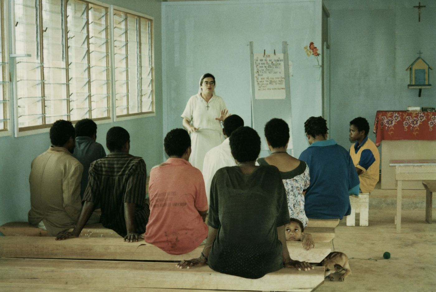 Colour photograph of a middle-aged nun, dressed in white. She is teaching a group of nine black adults in a rudimentary classroom. The students are sitting on wooden benches. A young boy and a dog are also present in the classroom.