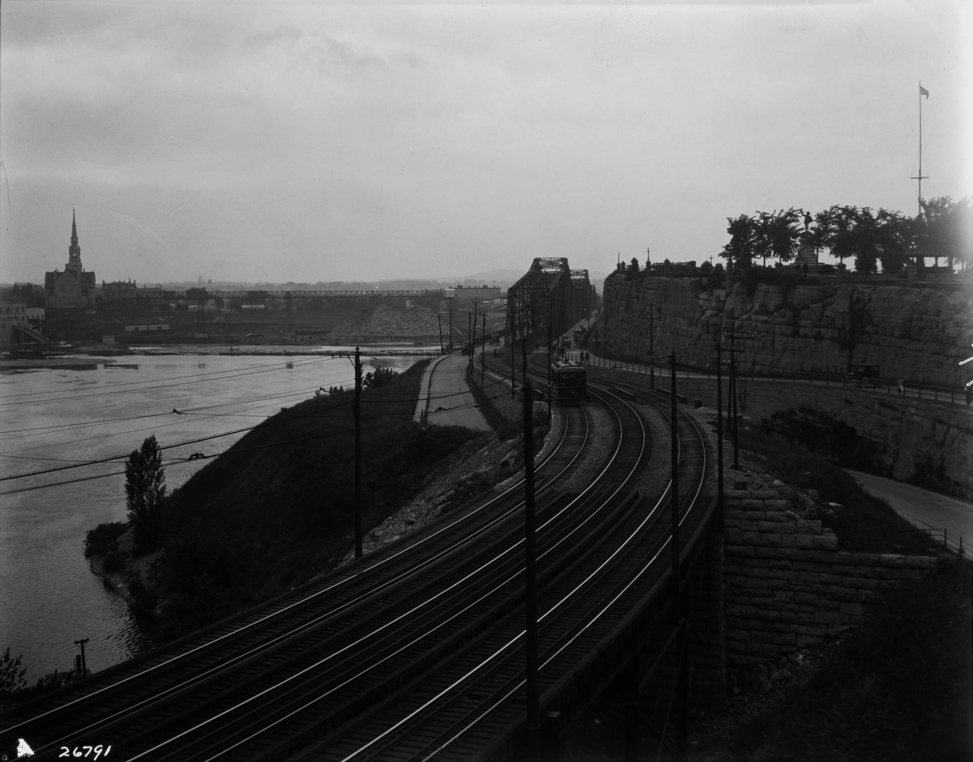Photographie en noir et blanc d’un paysage riverain. À l’avant-plan, trois voies de chemin de fer mènent à un pont de fer. De l’autre côté du cours d’eau, un paysage industriel dominé par une église imposante.