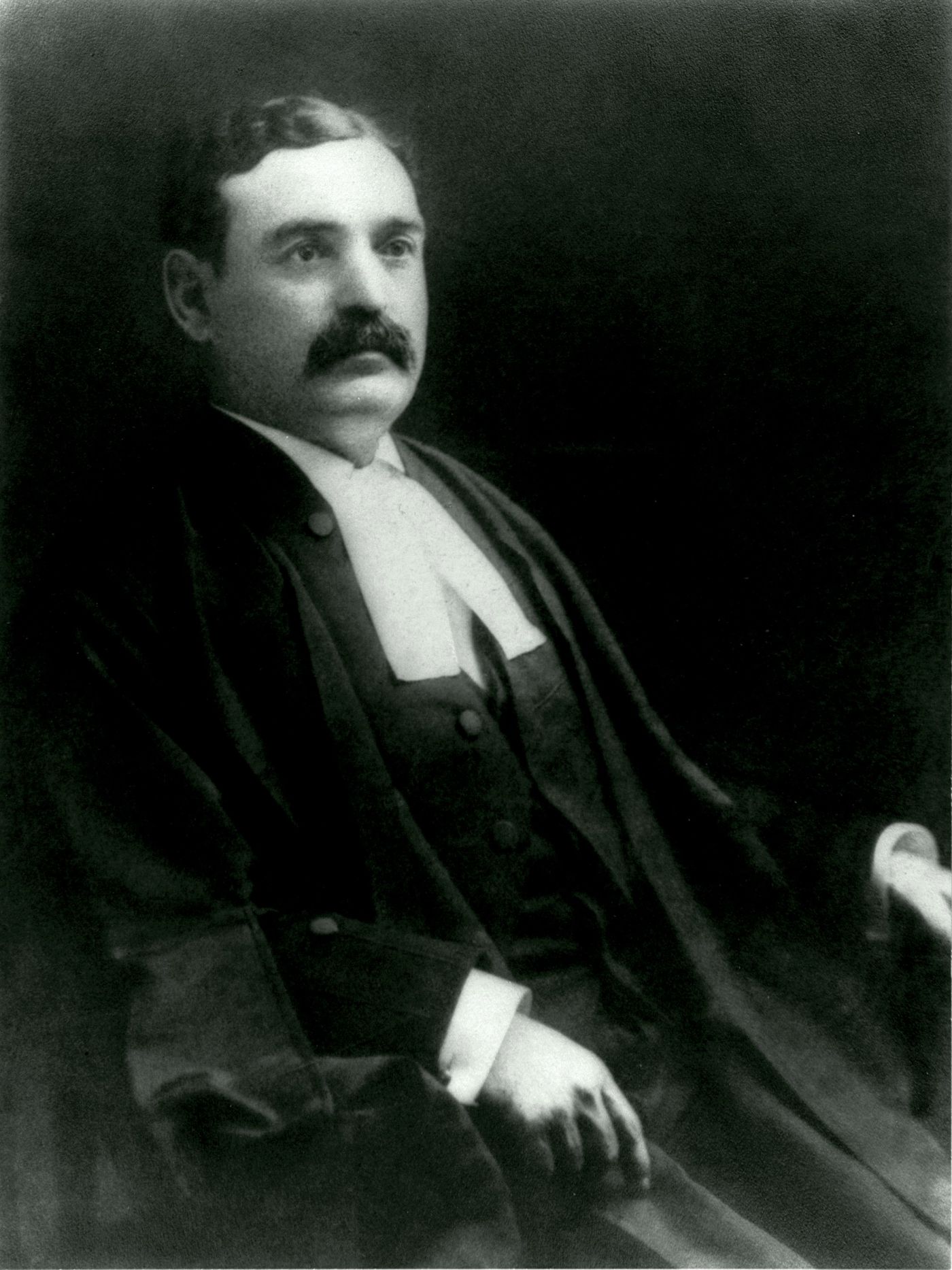 Black and white studio photograph of a middle-aged man sporting a mustache. He wears the long, loose gown and white collar of a lawyer.