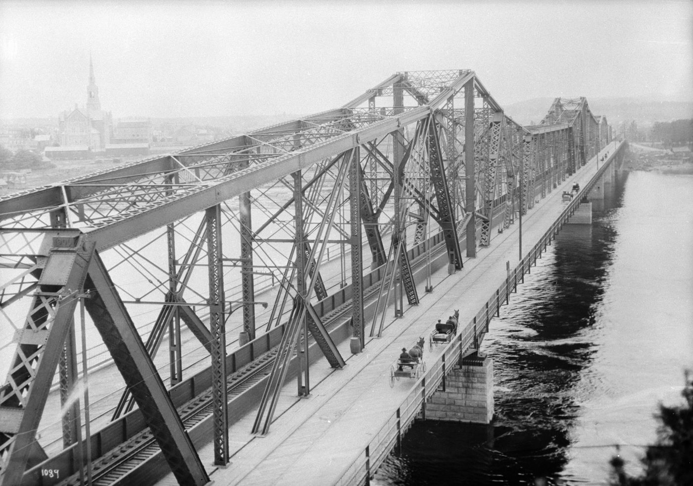 Photographie en noir et blanc d’un pont à treillis en acier surplombant une rivière. D’un côté du pont, des bogheis tirés par des chevaux se dirigent vers la rive où se trouve une église.