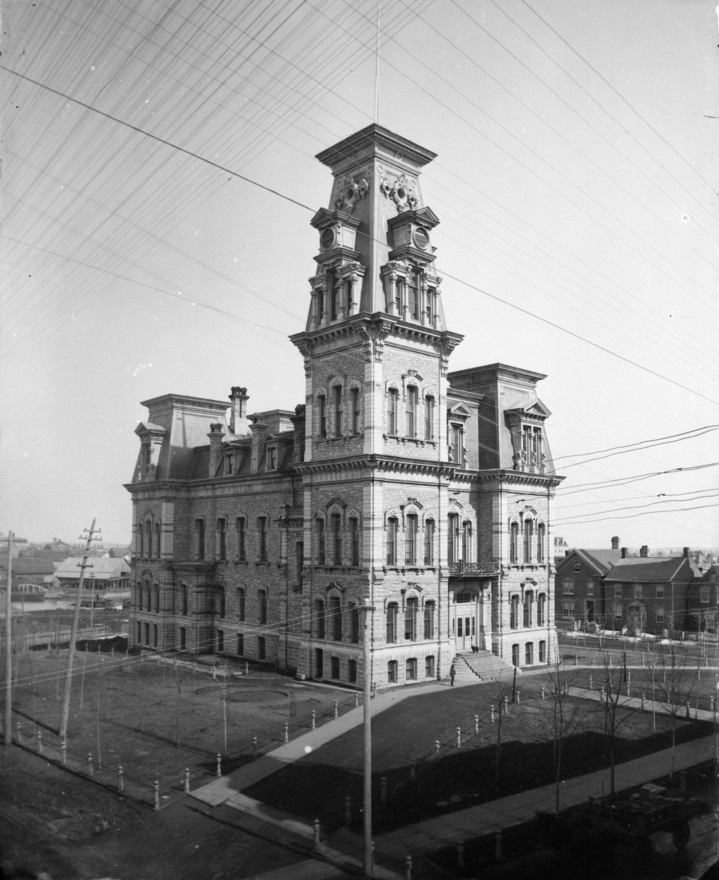 Photographie en noir et blanc d’un édifice en pierre à quatre étages, dominé par une tour à sa droite. L’édifice est séparé des maisons avoisinantes par un parc. Des lignes diagonales sont visibles en haut de l’image.