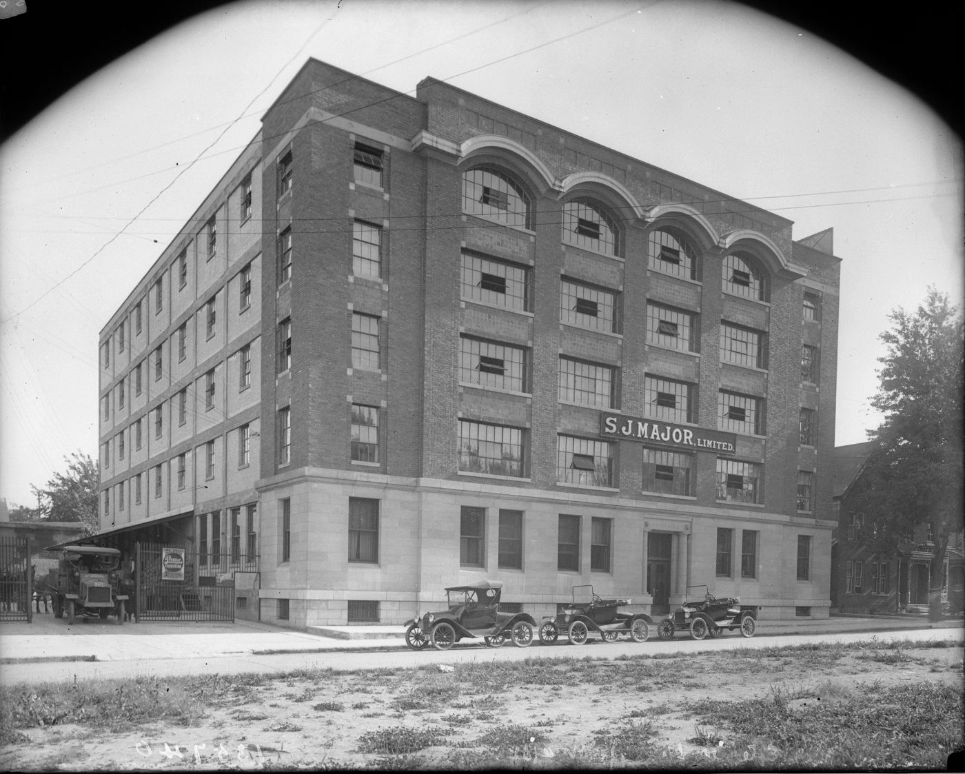 Black and white photograph of a six-storey brick building adorned with large windows. A sign bears the name of the business. Three vintage cars are parked in front of the main entrance.